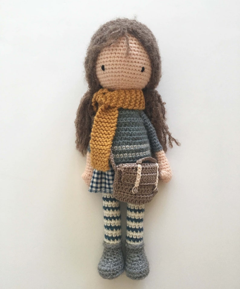 Peut inclure: Une poup&eacute;e en crochet avec des cheveux bruns, une &eacute;charpe jaune, un pull ray&eacute; gris et blanc, une jupe &agrave; carreaux, des leggings ray&eacute;s et des chaussures brunes. La poup&eacute;e tient un sac en crochet brun.