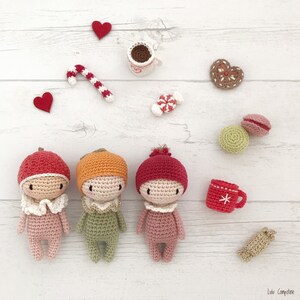 May include: Three crocheted dolls with different colored hats and outfits. The dolls are standing on a white wooden surface with various small decorative items around them.