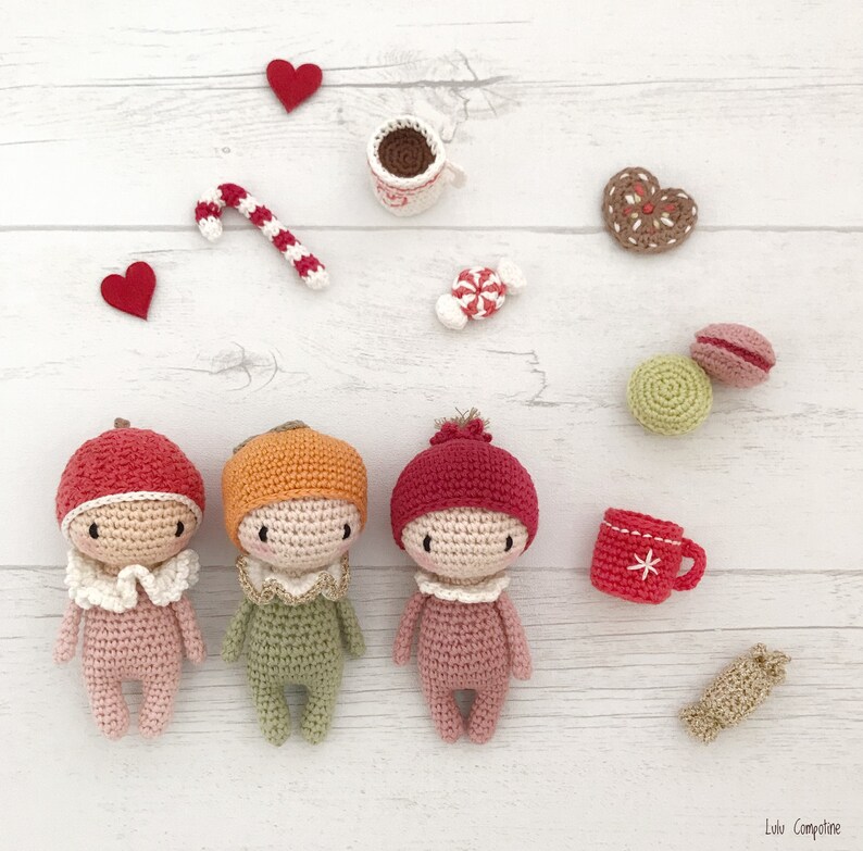 May include: Three crocheted dolls with different colored hats and outfits. The dolls are standing on a white wooden surface with various small decorative items around them.