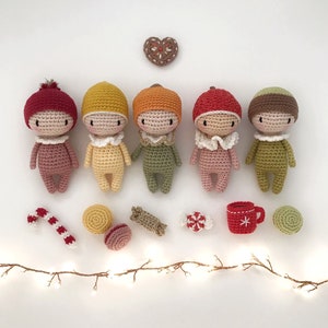 May include: Five crocheted dolls with different colored hats and outfits, including red, yellow, green, pink, and brown. The dolls are arranged in a row on a white surface with a string of lights, candy canes, and other decorations.