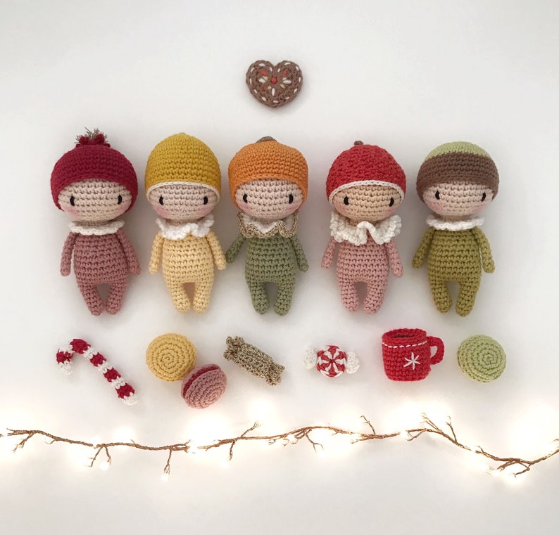 May include: Five crocheted dolls with different colored hats and outfits, including red, yellow, green, pink, and brown. The dolls are arranged in a row on a white surface with a string of lights, candy canes, and other decorations.