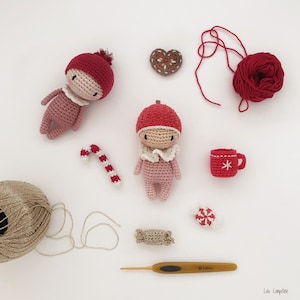 May include: Two crocheted dolls with red hats and pink outfits, a candy cane, a crochet heart, a crochet cup, a ball of yarn, a crochet candy, a crochet package, and a crochet hook.