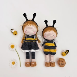 May include: Two handmade crochet dolls in bee costumes, featuring black and yellow striped outfits, black antennae, and small grey wings. Crochet bees, a daisy, and a honey pot are arranged around the dolls on a white background.
