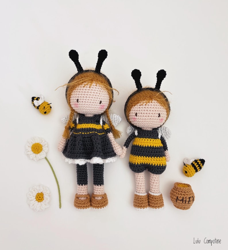 May include: Two handmade crochet dolls in bee costumes, featuring black and yellow striped outfits, black antennae, and small grey wings. Crochet bees, a daisy, and a honey pot are arranged around the dolls on a white background.