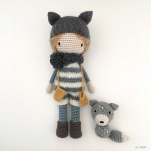 Op de afbeelding: Een gehaakte pop met een grijze muts met kattenoren, een gestreepte trui en een blauwe broek. De pop houdt gele wanten vast en staat naast een grijze gehaakte wolf.