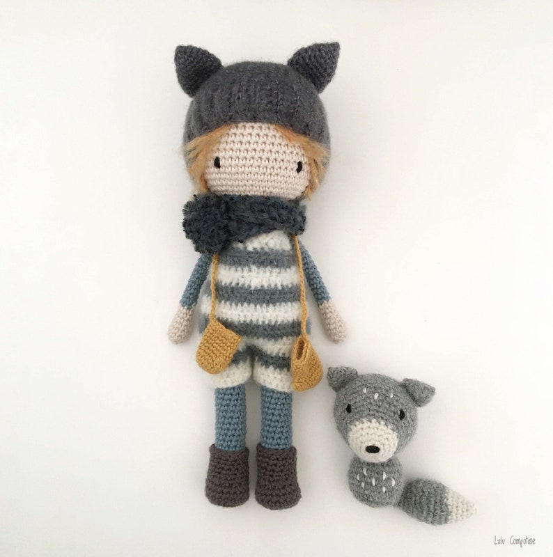 Op de afbeelding: Een gehaakte pop met een grijze muts met kattenoren, een gestreepte trui en een blauwe broek. De pop houdt gele wanten vast en staat naast een grijze gehaakte wolf.