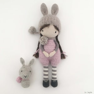 Pode incluir: Uma boneca de croch&ecirc; feita &agrave; m&atilde;o e um coelhinho. A boneca usa um macac&atilde;o rosa, um chap&eacute;u cinza com orelhas de coelho e meias listradas. O coelho tem um len&ccedil;o rosa. Ambos em um fundo branco.