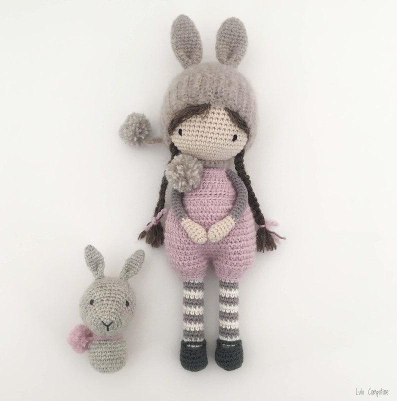 Pode incluir: Uma boneca de croch&ecirc; feita &agrave; m&atilde;o e um coelhinho. A boneca usa um macac&atilde;o rosa, um chap&eacute;u cinza com orelhas de coelho e meias listradas. O coelho tem um len&ccedil;o rosa. Ambos em um fundo branco.