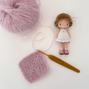May include: A crochet project featuring a ball of lavender yarn, a crochet hook, and a finished square. A small, handmade doll with brown hair and a white dress with pink accents is also present. The scene suggests a craft or hobby theme.