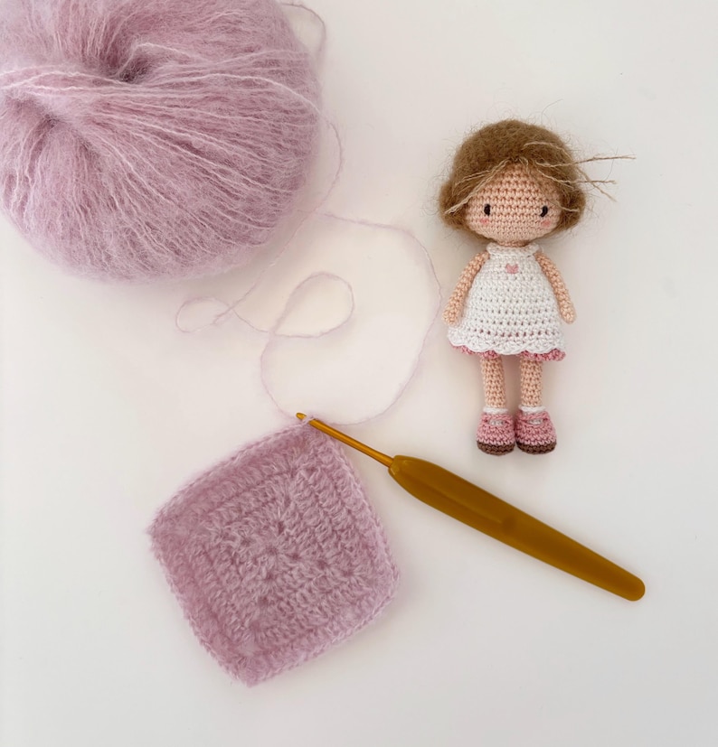 May include: A crochet project featuring a ball of lavender yarn, a crochet hook, and a finished square. A small, handmade doll with brown hair and a white dress with pink accents is also present. The scene suggests a craft or hobby theme.