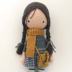 Peut inclure: Poup&eacute;e en crochet avec des cheveux bruns en tresses, portant une &eacute;charpe jaune, un pull bleu et gris, une jupe &agrave; carreaux et un sac jaune.