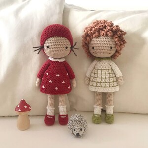 K&ouml;nnte beinhalten: Zwei geh&auml;kelte Puppen mit roten und wei&szlig;en Kleidern, braunen Haaren und schwarzen Augen. Die Puppen stehen auf einer wei&szlig;en Oberfl&auml;che mit einem kleinen Pilz und einem Igel.