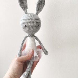 Puede incluir: Un conejito de crochet gris con un tut&uacute; blanco. El conejito tiene ojos negros y tiene los brazos extendidos.