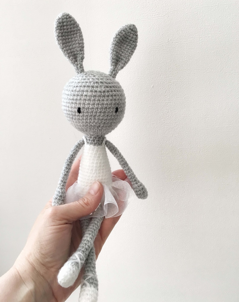 Puede incluir: Un conejito de crochet gris con un tut&uacute; blanco. El conejito tiene ojos negros y tiene los brazos extendidos.