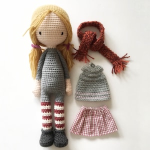 Peut inclure: Une poup&eacute;e en crochet aux cheveux blonds, portant une robe grise, des leggings ray&eacute;s rouge et blanc et des chaussures noires. La poup&eacute;e tient une &eacute;charpe rouge et a un haut en crochet gris et une jupe &agrave; carreaux rouge et blanc.