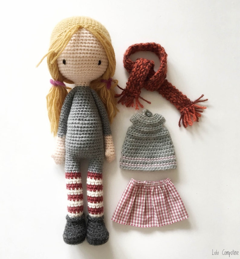 Peut inclure: Une poup&eacute;e en crochet aux cheveux blonds, portant une robe grise, des leggings ray&eacute;s rouge et blanc et des chaussures noires. La poup&eacute;e tient une &eacute;charpe rouge et a un haut en crochet gris et une jupe &agrave; carreaux rouge et blanc.