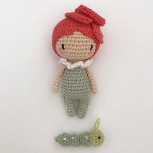 Puede incluir: Una mu&ntilde;eca de ganchillo hecha a mano con pelo rojo y un mono verde, y una peque&ntilde;a oruga verde. La mu&ntilde;eca tiene una cara beige con mejillas rosadas y un cuello blanco con volantes. La oruga es verde con puntos blancos y antenas amarillas.