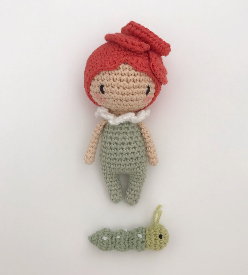 Puede incluir: Una mu&ntilde;eca de ganchillo hecha a mano con pelo rojo y un mono verde, y una peque&ntilde;a oruga verde. La mu&ntilde;eca tiene una cara beige con mejillas rosadas y un cuello blanco con volantes. La oruga es verde con puntos blancos y antenas amarillas.