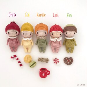 May include: Five crocheted dolls with different colored hats and outfits, including red, yellow, orange, pink, and green. The dolls are arranged in a row on a white surface. There are also several crocheted items, including a candy cane, a cookie, a macaron, a candy, and a heart-shaped cookie.