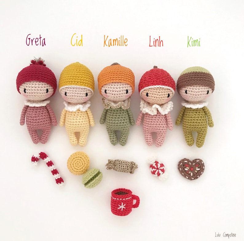May include: Five crocheted dolls with different colored hats and outfits, including red, yellow, orange, pink, and green. The dolls are arranged in a row on a white surface. There are also several crocheted items, including a candy cane, a cookie, a macaron, a candy, and a heart-shaped cookie.