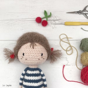 Puede incluir: Una mu&ntilde;eca de crochet con cabello casta&ntilde;o, cara rosa y una camisa a rayas azul y blanca. La mu&ntilde;eca lleva lazos rojos en el pelo. La mu&ntilde;eca est&aacute; rodeada de hilo y agujas de crochet.