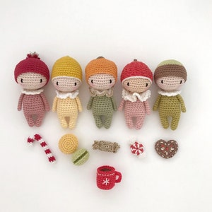 May include: Five crocheted amigurumi dolls in various colors, including red, yellow, orange, pink, and brown. The dolls are wearing different outfits and have smiling faces. The dolls are arranged in a row on a white background. There are also several small crocheted items, including a candy cane, a gingerbread cookie, and a cup of coffee.