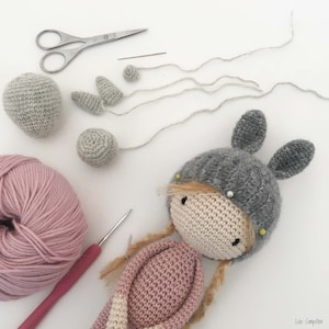 Pode incluir: Uma boneca de croch&ecirc; em processo de montagem, com um chap&eacute;u de coelho cinza e um vestido rosa. A imagem mostra v&aacute;rias pe&ccedil;as de croch&ecirc;, incluindo a cabe&ccedil;a, as orelhas e partes do corpo, juntamente com ferramentas como tesoura, uma agulha de croch&ecirc; e fio.