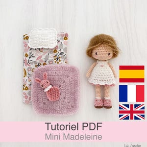 Peut inclure: Une poupée crochetée avec des cheveux bruns et une robe blanche, à côté d'un carré rose crocheté avec un appliqué de lapin et un tissu floral. L'image comprend également un petit oreiller crocheté blanc et le texte "Mini Madeleine".
