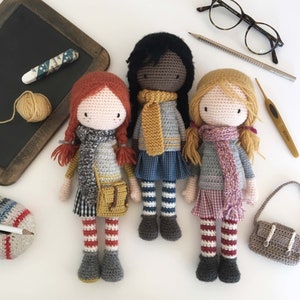 Peut inclure: Trois poup&eacute;es en crochet avec des tons de peau et des tenues diff&eacute;rentes. Les poup&eacute;es portent des &eacute;charpes, des robes et des collants ray&eacute;s. Les poup&eacute;es sont debout sur une surface blanche.