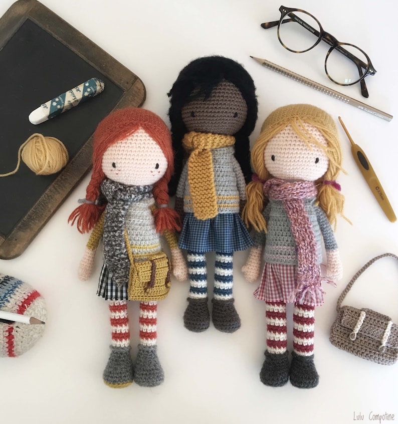 Peut inclure: Trois poup&eacute;es en crochet avec des tons de peau et des tenues diff&eacute;rentes. Les poup&eacute;es portent des &eacute;charpes, des robes et des collants ray&eacute;s. Les poup&eacute;es sont debout sur une surface blanche.