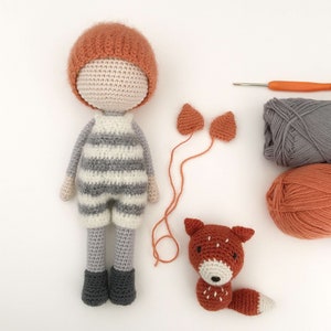 Op de afbeelding: Gebreide pop met een gestreepte outfit, een gebreide muts en een amigurumi vos. De pop is gemaakt van grijs en wit garen, en de muts is oranje. De vos is oranje en heeft een wit gezicht. Er zijn ook bollen garen op de achtergrond.