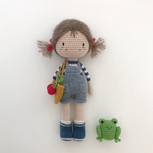 Puede incluir: Una mu&ntilde;eca de crochet con pelo marr&oacute;n, un mono azul y accesorios rojos y amarillos. La mu&ntilde;eca lleva una peque&ntilde;a bolsa de crochet. Una rana verde de crochet est&aacute; sentada delante de la mu&ntilde;eca.