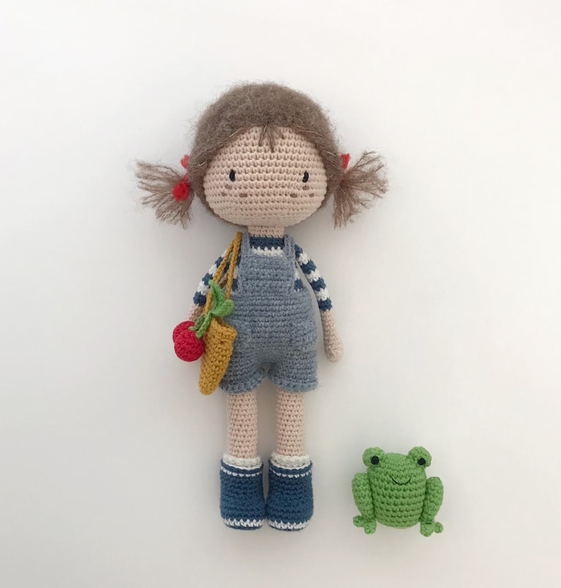 Puede incluir: Una mu&ntilde;eca de crochet con pelo marr&oacute;n, un mono azul y accesorios rojos y amarillos. La mu&ntilde;eca lleva una peque&ntilde;a bolsa de crochet. Una rana verde de crochet est&aacute; sentada delante de la mu&ntilde;eca.