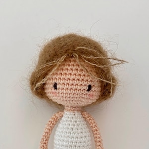 May include: A handmade crochet doll with light brown hair and a white dress. The doll's face is peach-colored with black eyes and rosy cheeks. The doll's arms and the top of the dress are a light peach color.