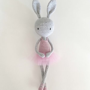 Puede incluir: Un conejito de crochet gris que lleva un vestido de crochet rosa y un tut&uacute; de tul. El conejito tiene ojos negros y est&aacute; sentado con las piernas cruzadas.