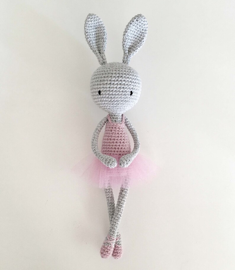Puede incluir: Un conejito de crochet gris que lleva un vestido de crochet rosa y un tut&uacute; de tul. El conejito tiene ojos negros y est&aacute; sentado con las piernas cruzadas.