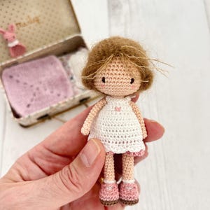 May include: A handmade crochet doll with brown hair, wearing a white dress with a pink trim and pink shoes. The doll has a sweet, simple face and is held in a hand. A small, decorated box is in the background.