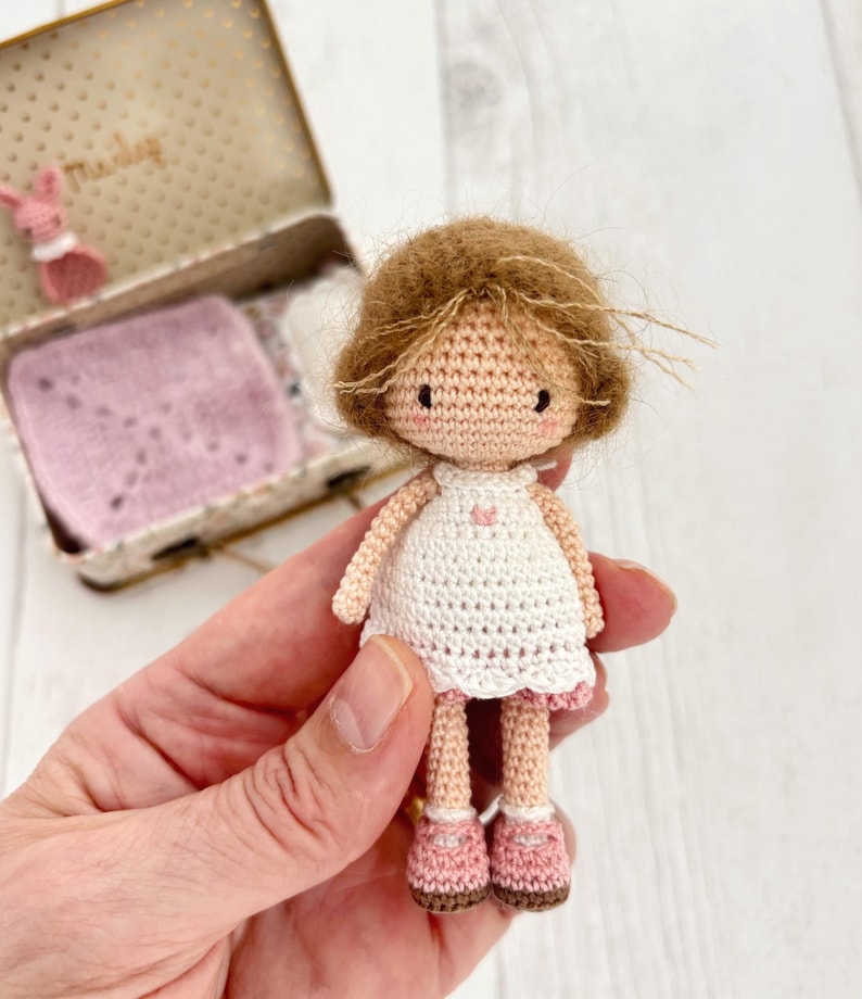 May include: A handmade crochet doll with brown hair, wearing a white dress with a pink trim and pink shoes. The doll has a sweet, simple face and is held in a hand. A small, decorated box is in the background.