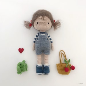 Puede incluir: Una mu&ntilde;eca de crochet con pelo marr&oacute;n, camisa a rayas azul y blanca, mono azul y zapatos rojos. La mu&ntilde;eca sostiene una peque&ntilde;a rana verde y una cesta con cerezas rojas.