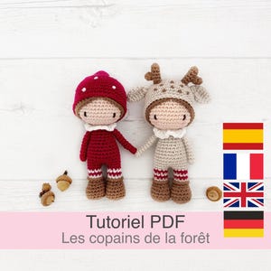 Może przedstawiać: Dwie szydełkowe lalki, jedna ubrana w grzyba, a druga w renifera, trzymają się za ręce. Lalki stoją na białym drewnianym podłożu. Tekst "Tutoriel PDF Les copains de la forêt" znajduje się na dole obrazu. Obraz zawiera również flagi Hiszpanii, Francji, Wielkiej Brytanii i Niemiec.