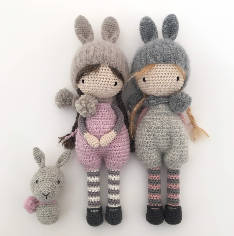 Pode incluir: Tr&ecirc;s bonecas de croch&ecirc; feitas &agrave; m&atilde;o e um coelhinho. As bonecas usam chap&eacute;us de orelhas de coelho e macac&otilde;es. Uma boneca tem uma roupa rosa, a outra cinza. O coelhinho tem um cachecol rosa. As bonecas t&ecirc;m tran&ccedil;as longas e pompons.