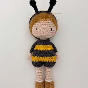 May include: A handmade crochet doll in a bee costume. The doll has a light brown complexion, rosy cheeks, and brown hair. It wears a black and yellow striped outfit with antennae. The doll's feet are brown with white trim.