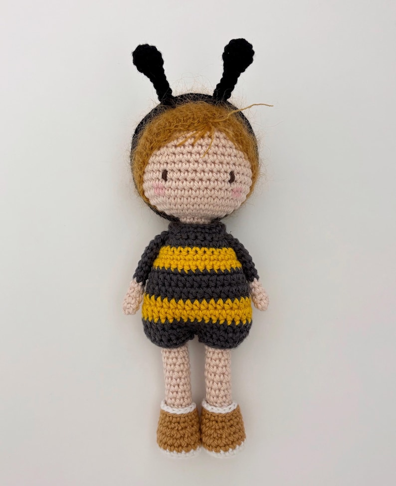 May include: A handmade crochet doll in a bee costume. The doll has a light brown complexion, rosy cheeks, and brown hair. It wears a black and yellow striped outfit with antennae. The doll's feet are brown with white trim.