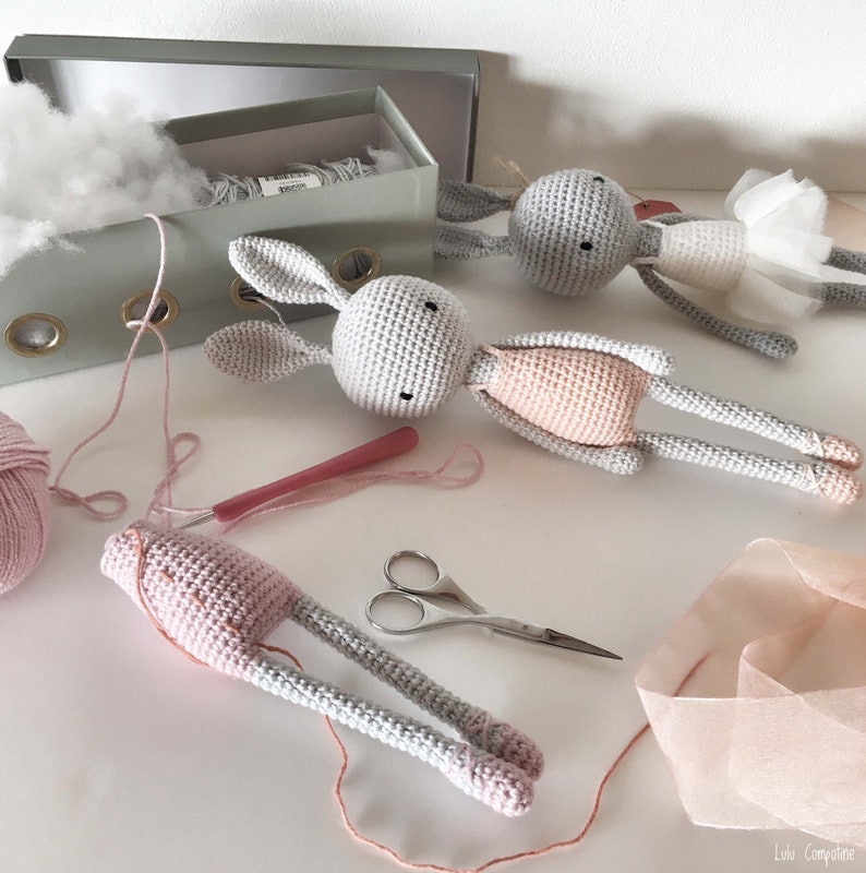 Puede incluir: Tres mu&ntilde;ecas de conejo de crochet gris con detalles rosas. Una conejita lleva un tut&uacute; rosa. Las mu&ntilde;ecas est&aacute;n sobre una superficie blanca con hilo rosa, un ganchillo rosa, unas tijeras y un trozo de tul rosa.
