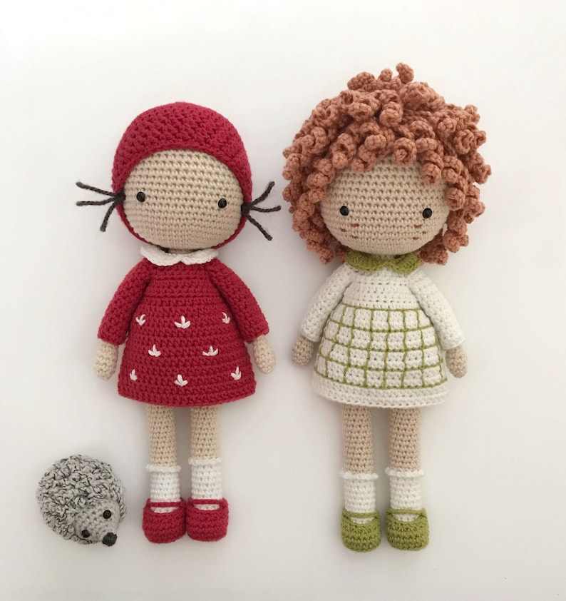 K&ouml;nnte beinhalten: Zwei geh&auml;kelte Puppen mit roten und wei&szlig;en Kleidern und braunen und gr&uuml;nen Haaren. Die Puppen stehen auf einer wei&szlig;en Oberfl&auml;che. Ein kleiner geh&auml;kelter Igel befindet sich im Vordergrund.