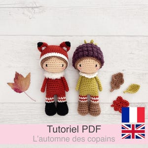Może przedstawiać: Dwie szydełkowe lalki, jedna przebrana za lisa w kolorze czerwono-czarnym, druga za żołądź w kolorze zielono-brązowym. Lalki znajdują się na białej powierzchni z jesiennymi liśćmi. Tekst "Tutoriel PDF L'automne des copains" znajduje się na dole.