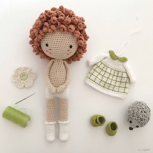 K&ouml;nnte beinhalten: Eine geh&auml;kelte Puppe mit braunen Locken, die ein wei&szlig;es Kleid und wei&szlig;e Socken tr&auml;gt. Die Puppe sitzt auf einer wei&szlig;en Oberfl&auml;che mit einem gr&uuml;nen Wollkn&auml;uel, einer Nadel, einer wei&szlig;en Blume, einem gr&uuml;nen Kleid und einem geh&auml;kelten Igel.