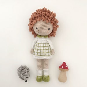 K&ouml;nnte beinhalten: Eine geh&auml;kelte Puppe mit roten Haaren, einem wei&szlig;en Kleid mit gr&uuml;nen Quadraten und wei&szlig;en Socken. Die Puppe steht neben einem geh&auml;kelten Igel und einem Holzpilz mit roten Punkten.