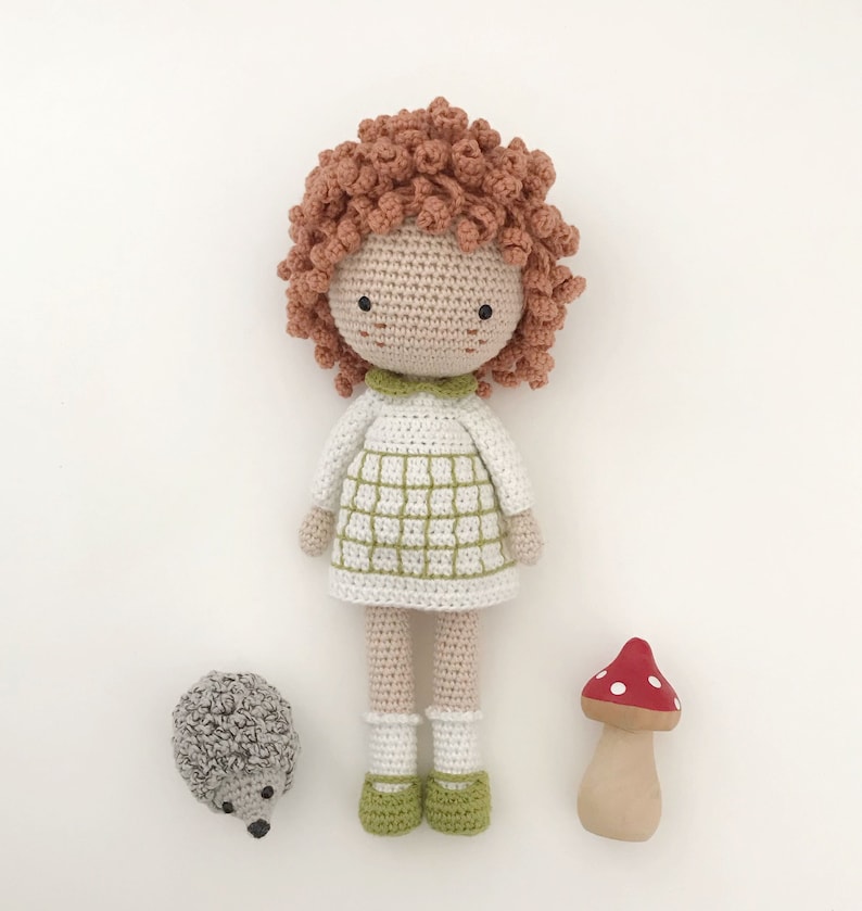 K&ouml;nnte beinhalten: Eine geh&auml;kelte Puppe mit roten Haaren, einem wei&szlig;en Kleid mit gr&uuml;nen Quadraten und wei&szlig;en Socken. Die Puppe steht neben einem geh&auml;kelten Igel und einem Holzpilz mit roten Punkten.