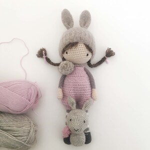 Pode incluir: Boneca de croch&ecirc; feita &agrave; m&atilde;o com um chap&eacute;u de coelho e um brinquedo de coelho a condizer. A boneca usa um vestido rosa e tem tran&ccedil;as castanhas com detalhes rosa. Meadas de l&atilde; cinza e rosa est&atilde;o no fundo.